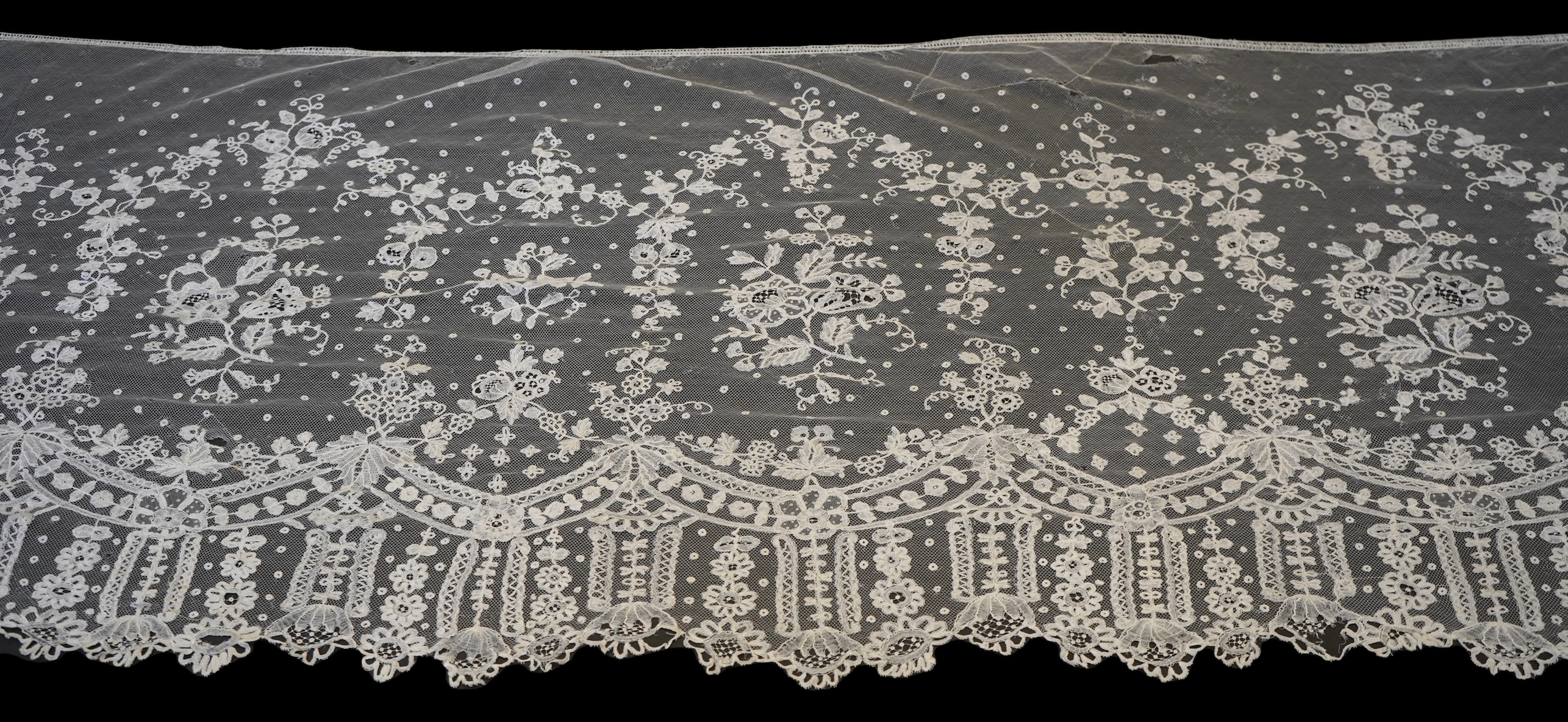 A deep elaborately designed flounce of mid 19th century Brussels bobbin appliquéd lace on net, with finished lace ends, worked with an intricate floral border below large floral cartouches framed in similar motifs and al